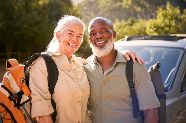 Happy couple with backpacks standing in front of a car. Couple posing with travel backpacks in front of a car