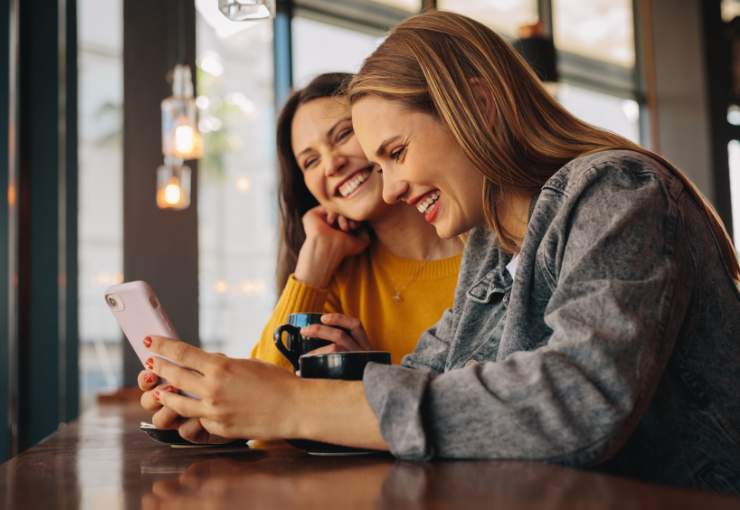 dos mujeres sonrientes con iPhones tomando café en una cafetería.